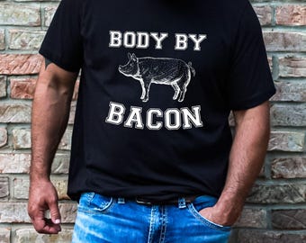 May include: Black t-shirt with the text "BODY BY BACON" above and below a detailed illustration of a pig. The shirt is worn by a person in blue jeans.
