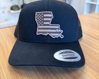 May include: Black snapback hat with a mesh back and a flat brim. The hat features a patch in the shape of Louisiana with a faded American flag design. The hat has a snapback closure and a Yupoong label.