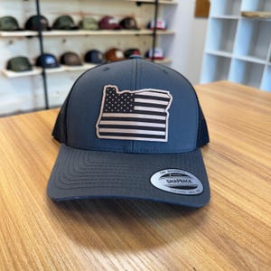 May include: A gray baseball cap with a black mesh back. The cap features a patch of the Oregon state outline with an American flag design. The brim has a curved shape, and the cap has a snapback closure.