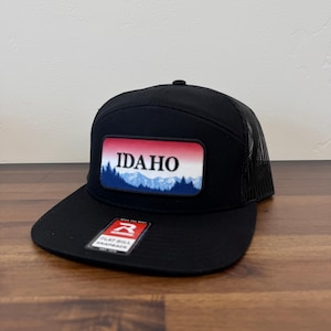May include: Black flat bill snapback hat with a mesh back. The front features a rectangular patch with the word "IDAHO" in white, above a mountain and tree graphic. The hat has a snap closure.