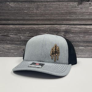 May include: A gray and black trucker hat with a wooden tree design patch. The hat features a gray front panel, a black mesh back, and a curved brim. The patch shows three trees.