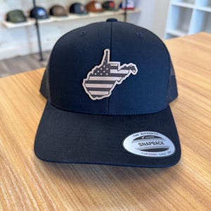 May include: Black snapback hat with a mesh back and a flat brim. The hat features a patch in the shape of West Virginia with an American flag design. The hat has a snap closure and a silver tag that reads "The Authentic Snapback".
