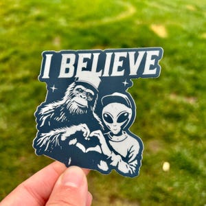 Puede incluir: Una pegatina azul marino con las palabras "I BELIEVE" en letras blancas. La pegatina presenta un gráfico de un Bigfoot y un extraterrestre haciendo una forma de corazón con sus manos. El fondo es verde.