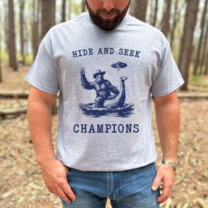 May include: A heather gray t-shirt with the words "HIDE AND SEEK CHAMPIONS" printed in navy blue. The graphic depicts Bigfoot, a UFO, and a Loch Ness Monster. The shirt is short-sleeved.