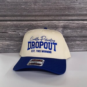 May include: A cream-colored baseball cap with a navy blue brim. The cap features the embroidered text "Gentle Parenting Dropout Est. This Morning" in navy blue. The hat has a curved brim and a button on top.