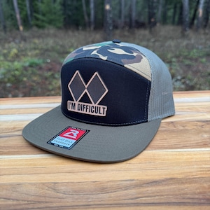 May include: A flat bill snapback hat with a black front panel featuring a copper-colored diamond design and the text "I'M DIFFICULT." The hat has a camouflage pattern on the top, a gray mesh back, and a green brim.