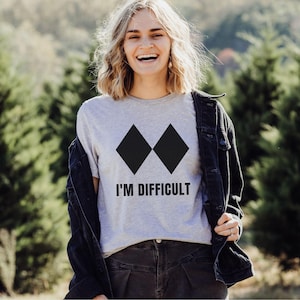 May include: A light gray t-shirt with two black diamond shapes and the text "I'M DIFFICULT" in black. The shirt is being worn with a black jacket. The background is a blurred view of green trees.