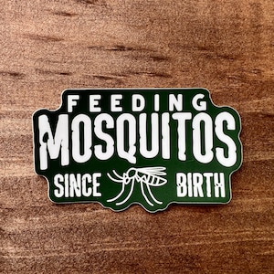 May include: A green sticker with white text that reads "FEEDING MOSQUITOS SINCE BIRTH." A white graphic of a mosquito is below the word "SINCE." The sticker is on a brown wooden surface.