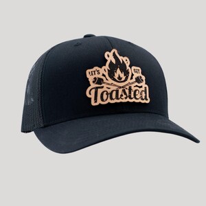 May include: Black trucker hat with a mesh back and a curved brim. The hat features a brown leather patch with the words "Let's Get Toasted" and a graphic of a campfire with marshmallows.