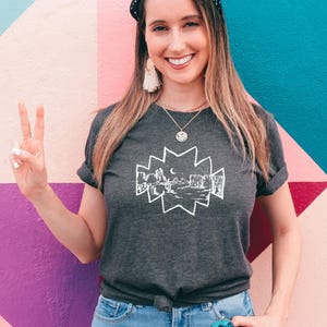May include: Dark gray t-shirt featuring a white graphic of a desert landscape with cacti, mountains, and a crescent moon. The shirt has rolled-up sleeves and a knotted front. The background is a colorful wall with pink, purple, and teal sections.
