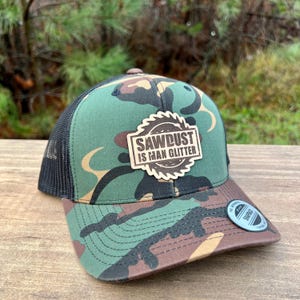 May include: A camouflage trucker hat with a black mesh back and a curved brim. The front panel features a patch with the text "SAWDUST IS MAN GLITTER" in a wood-grain design. The hat has a snapback closure and a small circular tag on the brim.