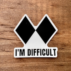 May include: A white sticker with a black and white diamond design, and the text "I'M DIFFICULT" in black. The sticker is on a wooden surface.