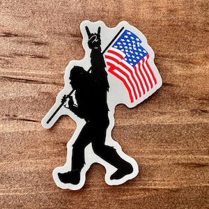 May include: A sticker featuring a black silhouette of Bigfoot holding an American flag. The flag has red and white stripes and a blue field with white stars. Bigfoot is making a rock and roll hand gesture.