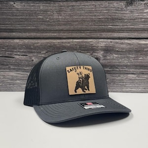 May include: Charcoal gray trucker hat with a black mesh back and a curved brim. A square, tan leather patch features a graphic of a person riding a bear with the text "SAFETY THIRD".