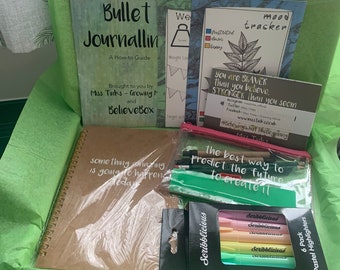May include: A green gift box filled with stationery supplies, including a brown notebook with the text "something amazing is going to happen today", a green pencil case with colorful pens and pencils, and a pack of pastel highlighters. The box also includes a guide to bullet journaling and motivational posters.