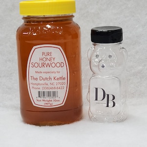 May include: A glass jar of pure honey labeled "Sourwood" with a yellow lid. The honey is made by The Dutch Kettle in Hamptonville, NC. The jar is next to a clear plastic bear-shaped honey dispenser with a black lid and the letters "DB" printed on the front.