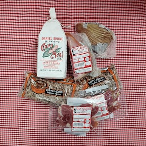 May include: A red and white checkered tablecloth with a variety of food items, including a bag of Daniel Boone Self-Rising Corn Meal Mix, a package of pinto beans, a package of country ham, and a package of sliced ham.