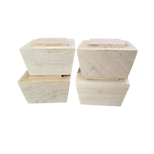 May include: Four unfinished wooden furniture feet. Each foot is a light-colored, square block with a slightly tapered design. The top of each foot has a smaller, rectangular piece attached.