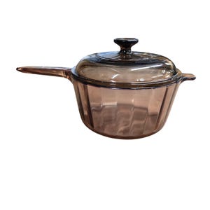 May include: A translucent amber-colored glass saucepan featuring a ribbed, vertical fluted design on the exterior body. The cookware includes a matching glass lid with a central knob handle and a single elongated glass handle on the side.