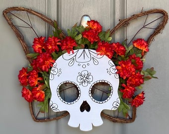 May include: A white sugar skull wreath with a black and white design. The wreath is decorated with red and green artificial flowers.