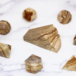 Puede incluir: Un conjunto de siete dados de piedra de diferentes formas y tamaños, con un patrón de mármol marrón y blanco. Los dados están numerados del 1 al 6, y el conjunto también incluye un d8 y un d20.