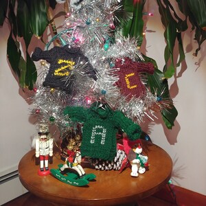 May include: A silver tinsel Christmas tree decorated with four knitted sweater ornaments, each with a different letter, spelling out the word "HOME". The tree is on a wooden table with a nutcracker, a rocking horse, and a small stuffed animal.