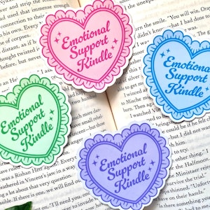 K&ouml;nnte beinhalten: Vier herzf&ouml;rmige Aufkleber mit dem Text "Emotional Support Kindle" in einer verspielten Schriftart. Jeder Aufkleber hat einen gewellten Rand und ist in einer anderen Pastellfarbe gehalten: Rosa, Gr&uuml;n, Blau und Lila. Auf einem offenen Buch.