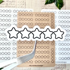 May include: A collection of star-shaped stickers in various sizes and colors. The stickers are arranged on white and kraft paper sheets. A large sticker with five stars is held by tweezers. The text "#ByJulieMarieDesigns" is visible.