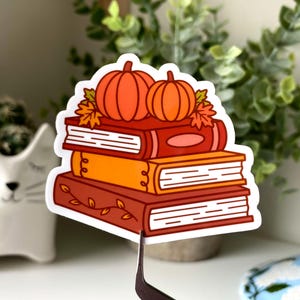 May include: A sticker featuring a stack of books with two orange pumpkins and fall leaves on top. The books are in shades of orange and brown. The sticker is held by tweezers.
