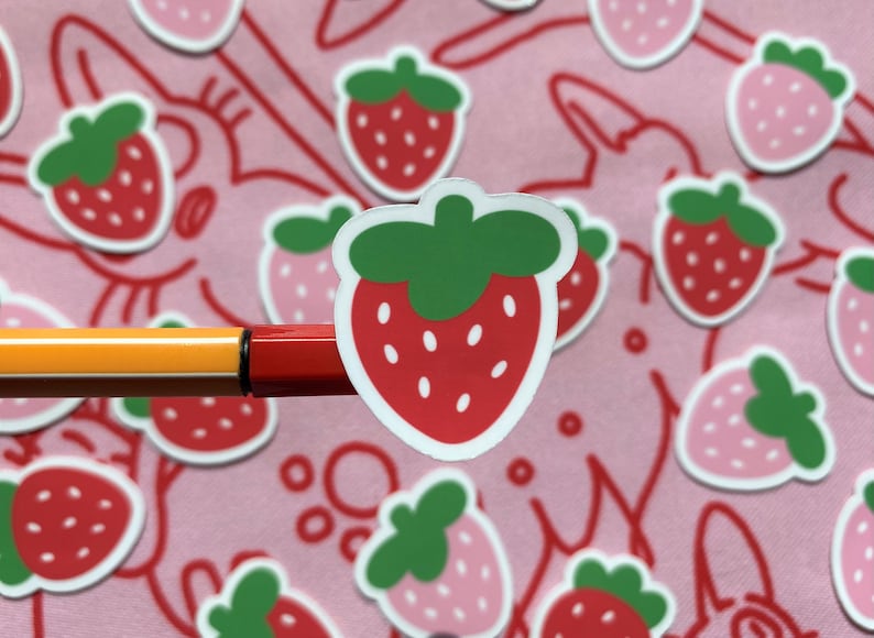 May include: A close-up of a red and green strawberry sticker on a pink background. The sticker is surrounded by other strawberry stickers in various colors.
