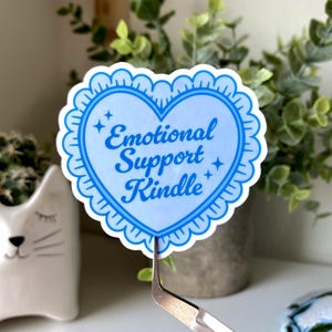 K&ouml;nnte beinhalten: Ein hellblauer herzf&ouml;rmiger Aufkleber mit den Worten "Emotional Support Kindle" in einer Schreibschrift. Der Aufkleber hat einen gewellten Rand und wird von einer Pinzette gehalten. Der Hintergrund umfasst einen katzenf&ouml;rmigen Pflanztopf und Gr&uuml;n.