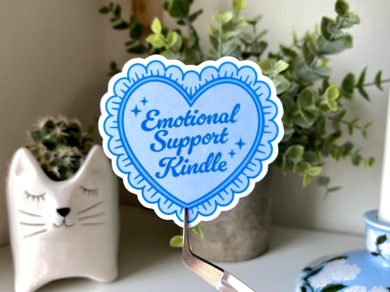 K&ouml;nnte beinhalten: Ein hellblauer herzf&ouml;rmiger Aufkleber mit den Worten "Emotional Support Kindle" in einer Schreibschrift. Der Aufkleber hat einen gewellten Rand und wird von einer Pinzette gehalten. Der Hintergrund umfasst einen katzenf&ouml;rmigen Pflanztopf und Gr&uuml;n.