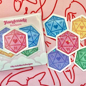 Pode incluir: Um conjunto de seis adesivos hologr&aacute;ficos iridescentes com um design em forma de cora&ccedil;&atilde;o dentro de um dado de 20 faces. Os adesivos s&atilde;o de v&aacute;rias cores, incluindo rosa, azul, amarelo, verde e roxo. Os adesivos est&atilde;o sobre um fundo branco com um design de nuvens rosa e branco. O texto "Handmade Stickers" e "@ByJulieMarieDesigns" est&aacute; impresso no fundo.