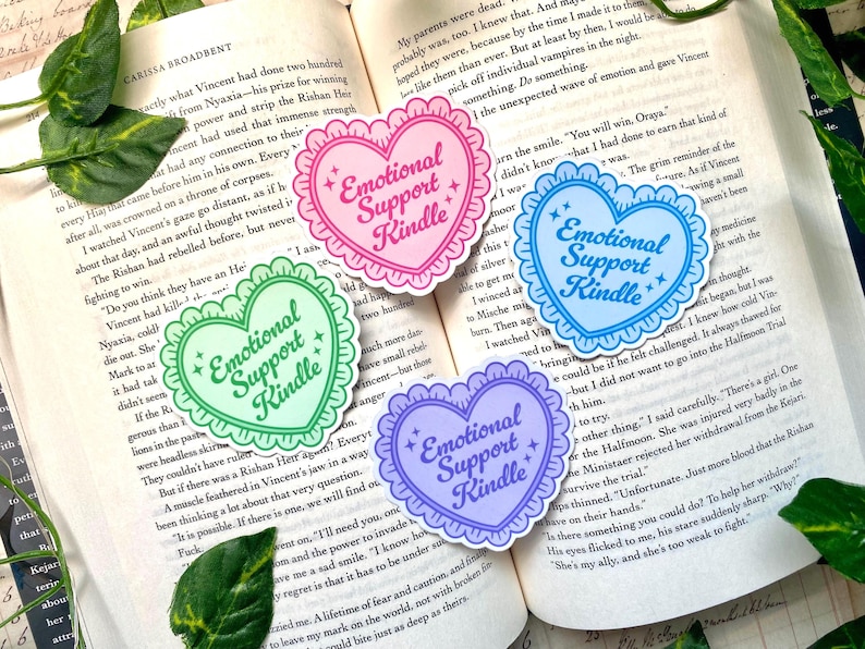 K&ouml;nnte beinhalten: Vier herzf&ouml;rmige Aufkleber mit dem Text "Emotional Support Kindle" in einer verspielten Schriftart. Jeder Aufkleber hat einen gewellten Rand und ist in einer anderen Pastellfarbe gehalten: Rosa, Gr&uuml;n, Blau und Lila. Auf einem offenen Buch.
