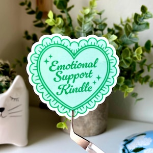 K&ouml;nnte beinhalten: Ein herzf&ouml;rmiger Aufkleber mit den Worten "Emotional Support Kindle" in gr&uuml;ner Schrift. Der Aufkleber hat einen hellgr&uuml;nen Hintergrund und einen gewellten Rand. Der Aufkleber wird von einer Pinzette gehalten.