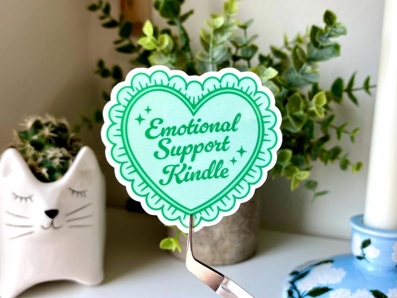 K&ouml;nnte beinhalten: Ein herzf&ouml;rmiger Aufkleber mit den Worten "Emotional Support Kindle" in gr&uuml;ner Schrift. Der Aufkleber hat einen hellgr&uuml;nen Hintergrund und einen gewellten Rand. Der Aufkleber wird von einer Pinzette gehalten.