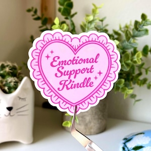 K&ouml;nnte beinhalten: Ein rosa herzf&ouml;rmiger Aufkleber mit dem Text "Emotional Support Kindle". Der Aufkleber hat einen gewellten Rand und Sternakzente. Im Hintergrund befindet sich ein wei&szlig;er Katzen-Pflanztopf mit einem Kaktus.