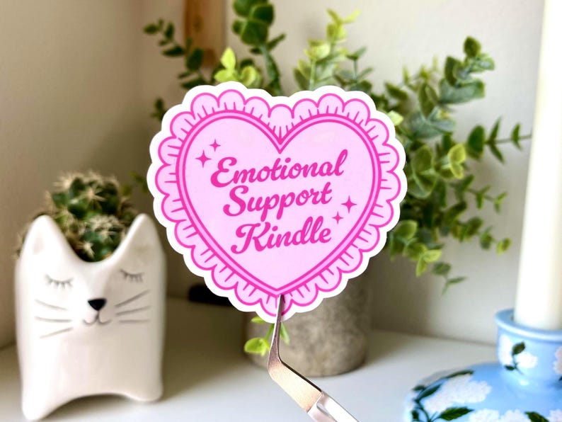 K&ouml;nnte beinhalten: Ein rosa herzf&ouml;rmiger Aufkleber mit dem Text "Emotional Support Kindle". Der Aufkleber hat einen gewellten Rand und Sternakzente. Im Hintergrund befindet sich ein wei&szlig;er Katzen-Pflanztopf mit einem Kaktus.