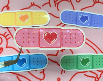 May include: Five colorful band-aid stickers with a heart design. The band-aids are yellow, pink, blue, green, and light blue. Each band-aid has a different colored heart in the center.