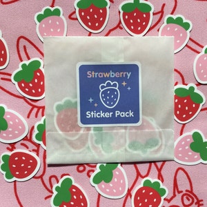 May include: A white envelope with a sticker pack of strawberry-themed stickers. The envelope has a sticker on the front that says "Strawberry Sticker Pack". The background is pink with a pattern of strawberries.