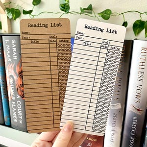 May include: Two brown and white reading list bookmarks with a black border and text. The bookmarks have spaces to write the name of the book, author, year, goal, date, and rating. The bookmarks are on a bookshelf with books behind them.