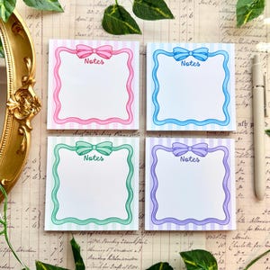 May include: Four square sticky note pads with a striped background and a wavy border featuring a bow at the top. Each pad has the word Notes printed in the center. The pads are pink, blue, green, and purple, arranged in a two-by-two grid layout.