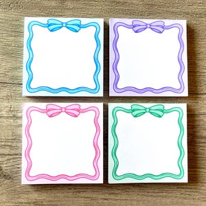 May include: Four blank notepads with decorative borders. Each notepad has a different colored border and a bow in the center. The colors are blue, purple, pink, and green.