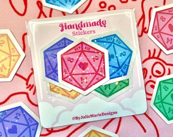 May include: Five iridescent holographic stickers of 20-sided dice with a heart in the center. The dice are pink, blue, green, and two shades of yellow. The stickers are arranged on a white card with the text "Handmade Stickers" and "@ByJulieMarieDesigns".