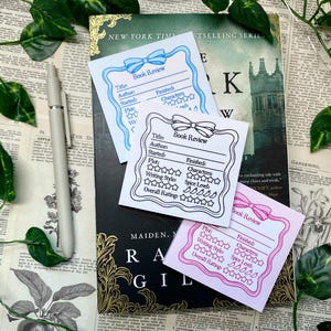 May include: Three printable book review cards with a black and white outline and a bow design. The cards are blue, pink, and white. The cards have sections for title, author, started, finished, plot, characters, writing style, spice level, and overall rating.