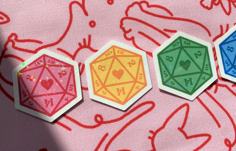 Pode incluir: Quatro adesivos iridescentes de um dado de 20 faces com um cora&ccedil;&atilde;o no centro. Os dados s&atilde;o de cor rosa, amarelo, verde e azul.