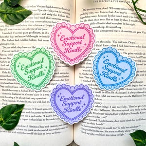 K&ouml;nnte beinhalten: Vier herzf&ouml;rmige Aufkleber mit dem Text "Emotional Support Kindle". Jeder Aufkleber hat einen gewellten Rand und ist in einer anderen Pastellfarbe gehalten: Gr&uuml;n, Rosa, Blau und Lila. Sie liegen auf einem offenen Buch.