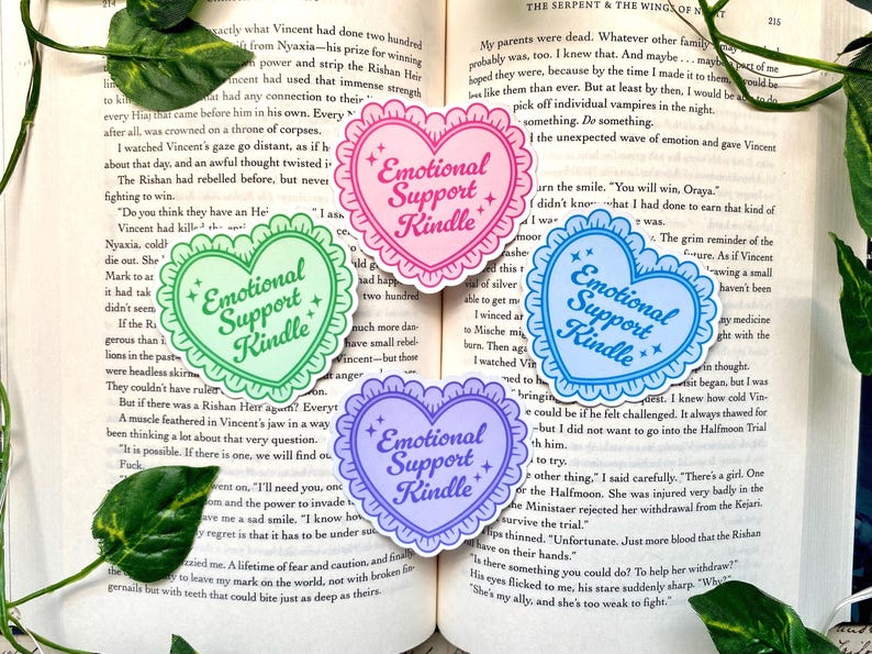 K&ouml;nnte beinhalten: Vier herzf&ouml;rmige Aufkleber mit dem Text "Emotional Support Kindle". Jeder Aufkleber hat einen gewellten Rand und ist in einer anderen Pastellfarbe gehalten: Gr&uuml;n, Rosa, Blau und Lila. Sie liegen auf einem offenen Buch.