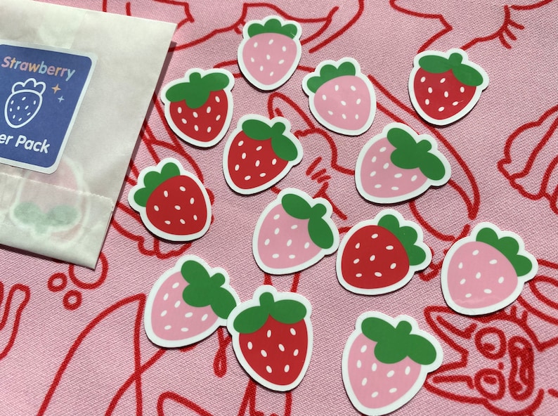 May include: A sheet of 12 strawberry stickers. The stickers are in two colors: pink and red. The stickers are on a pink background with red lines.