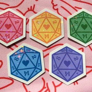 Pode incluir: Cinco adesivos coloridos de um dado de 20 faces com um cora&ccedil;&atilde;o no centro. Os dados s&atilde;o rosa, amarelo, verde, azul e roxo.